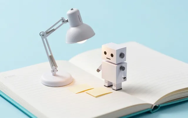 A small white paper robot compares two paper slips under a desk lamp beside an open ledger